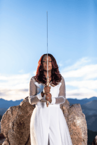 Lacricia Darling raising a gold sword above her head on a mountain overlook, symbolizing faith and courage